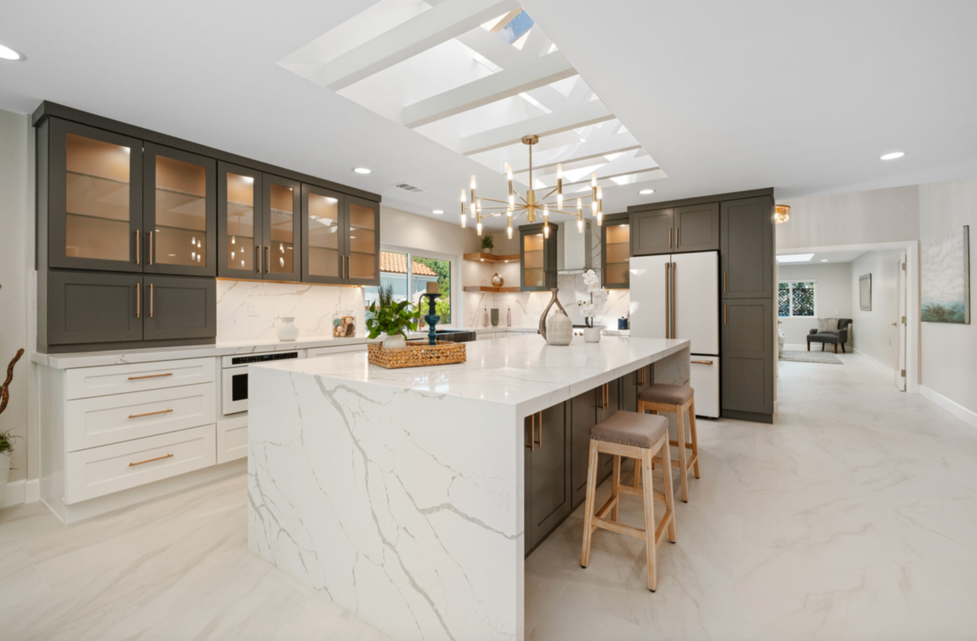 Skylit kitchen with waterfall marble island, brass chandelier, and glass-front cabinetry